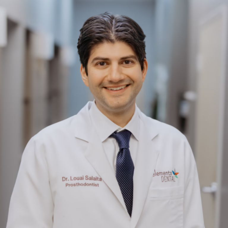 Smiling male dentist in a white coat, name tag reads Dr. Louai Salaita, Prosthodontist, in a dental clinic hallway at Elements Dental.