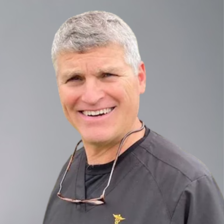 Smiling middle-aged man with short gray hair wearing dark scrubs and glasses hanging from his neck against a neutral background.