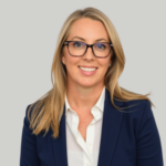 Professional headshot of a smiling blonde woman wearing glasses and a navy blazer against a light gray background, suitable for a corporate profile.