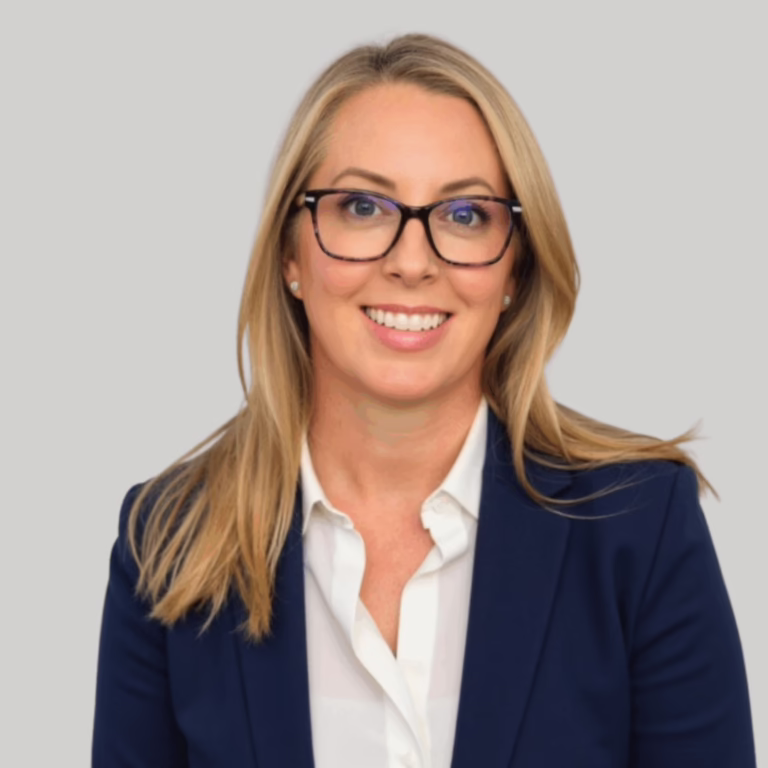 Professional headshot of a smiling blonde woman wearing glasses and a navy blazer against a light gray background, suitable for a corporate profile.