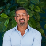 Smiling man with short dark hair and goatee in a light blue button-down shirt, standing before lush green tropical plants.