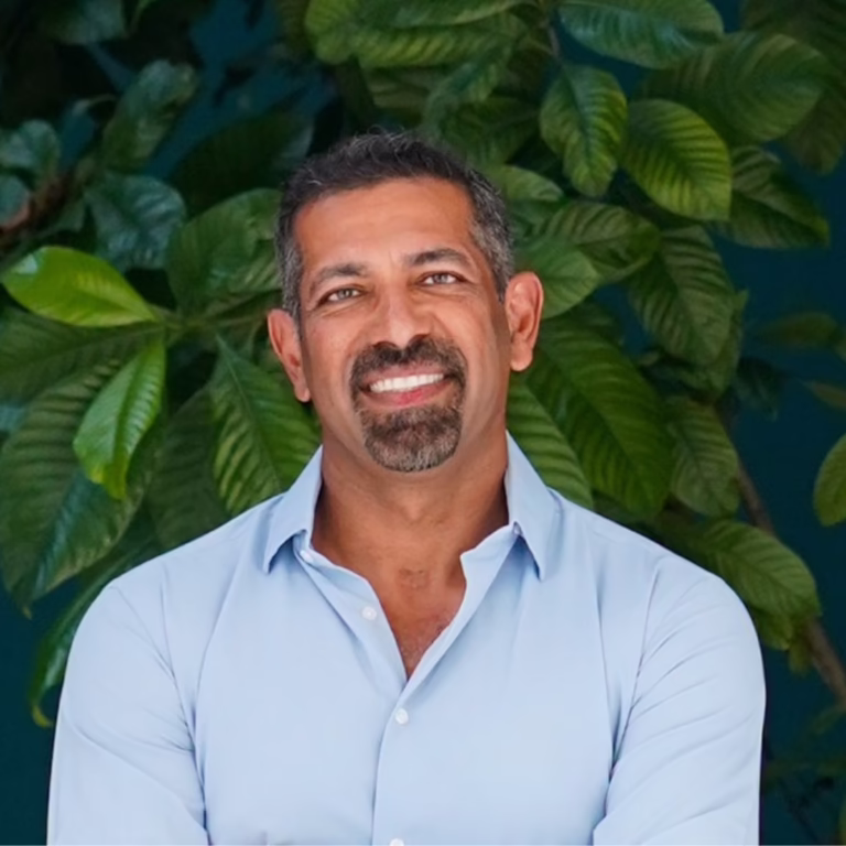 Smiling man with short dark hair and goatee in a light blue button-down shirt, standing before lush green tropical plants.