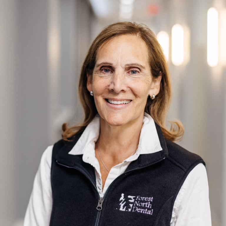 Smiling middle-aged woman wearing a Forest North Dental vest over a white shirt, standing in a hallway.