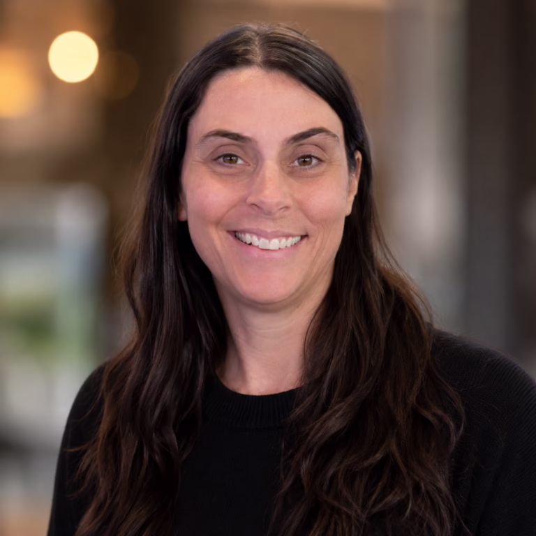 Smiling woman with long dark hair wearing a black sweater, posed for a portrait in a softly blurred office scene
