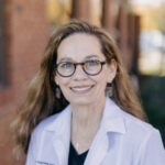 Smiling woman in a white lab coat and round glasses, outdoors, facing the camera.
