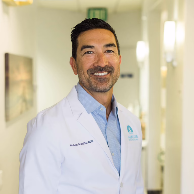 Smiling male dentist in a white coat standing in a bright hallway, name badge reads Dr. Robert Schaffer DDS.