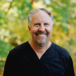 Smiling man with gray hair and goatee wearing black medical scrubs, outdoors with a blurred green-yellow background.