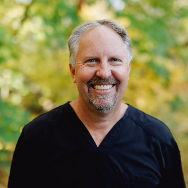 Smiling man with gray hair and goatee wearing black medical scrubs, outdoors with a blurred green-yellow background.