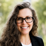 Smiling woman with long wavy hair and black glasses, wearing a black blazer outdoors.