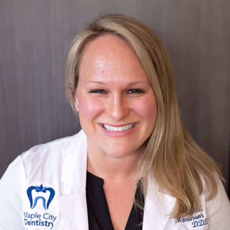 Smiling woman in a white dental coat with Maple City Dentistry logo on the chest, portrait against a dark background.