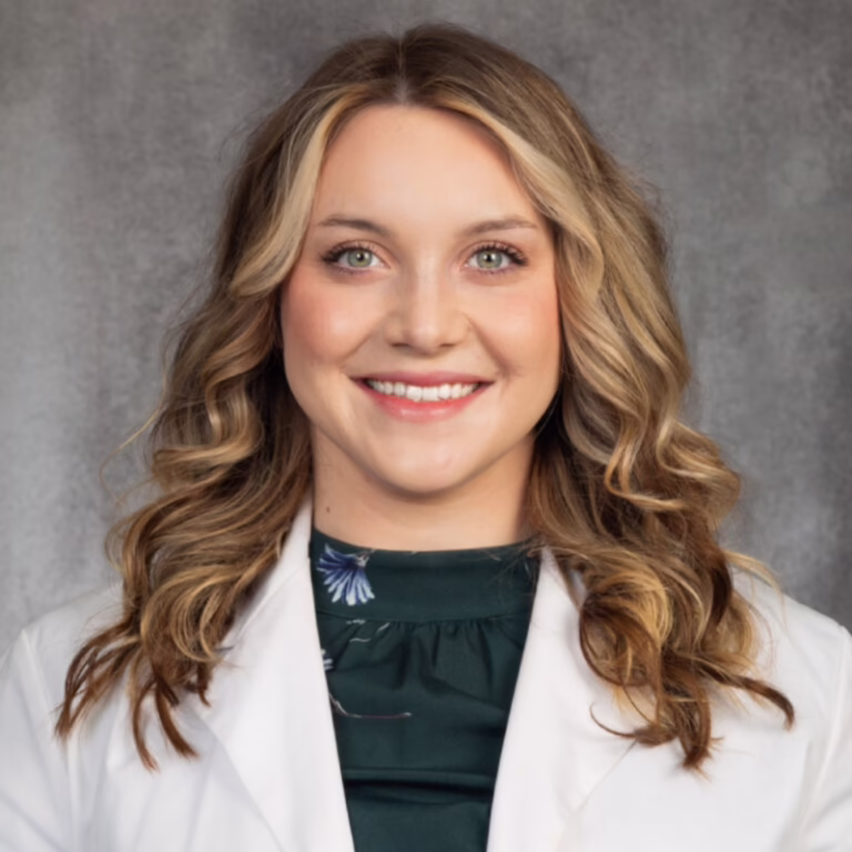Professional headshot: smiling woman with wavy light-brown hair, wearing a white lab coat.