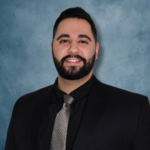 Professional headshot of a man with dark hair and beard, wearing a black suit and gray patterned tie against a blue backdrop.