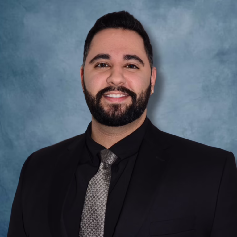 Professional headshot of a man with dark hair and beard, wearing a black suit and gray patterned tie against a blue backdrop.