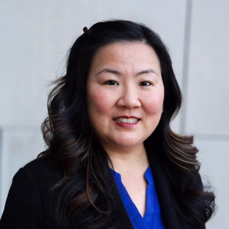 Portrait of a smiling Asian woman with long dark hair, wearing a blue blouse and black blazer in a neutral setting.