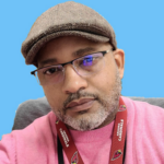 Close-up portrait of a man with a gray beard, wearing a brown flat cap, glasses, and a pink sweater against a blue background. (informative)