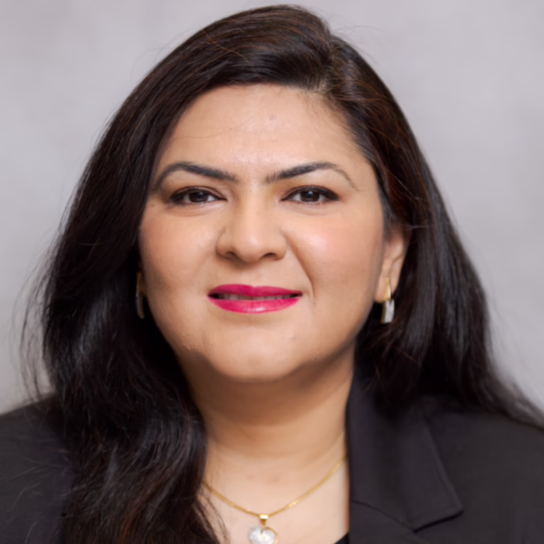 Head-and-shoulders portrait of a smiling woman with dark hair in a black blazer, wearing gold jewelry, against a gray backdrop.