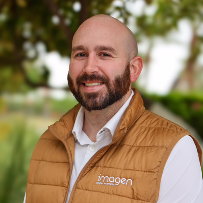 Smiling man with a beard and shaved head wearing a tan puffer vest over a white shirt; outdoors, vest shows a logo.