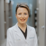 Female dentist in a white coat standing in a clinic hallway, smiling at the camera.