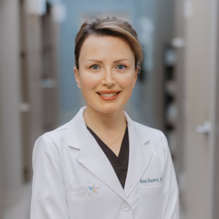 Female dentist in a white coat standing in a clinic hallway, smiling at the camera.