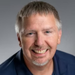 Portrait of a smiling middle-aged man with short gray hair and a trimmed goatee, wearing a navy shirt against a gray background.