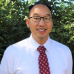 Smiling man with glasses in a white dress shirt and red patterned tie, outdoors with green foliage behind him.