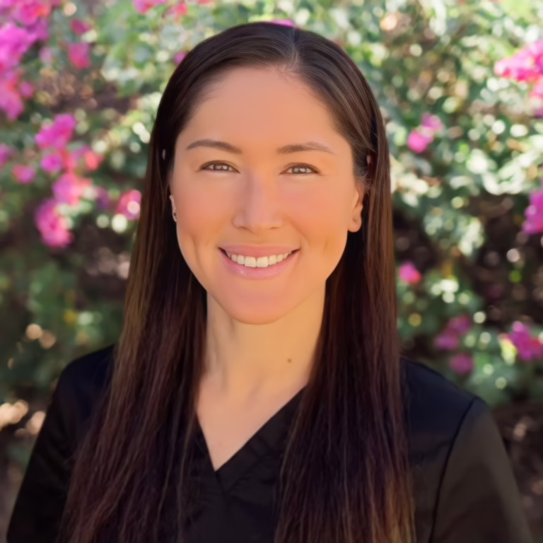 Portrait of a smiling woman with long dark hair wearing a black top, outdoors with pink flowers in the background.