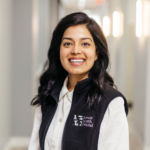 Smiling woman with dark hair in a Forest North Dental vest, standing in a bright indoor hallway.