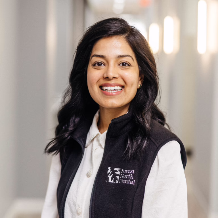 Smiling woman with dark hair in a Forest North Dental vest, standing in a bright indoor hallway.