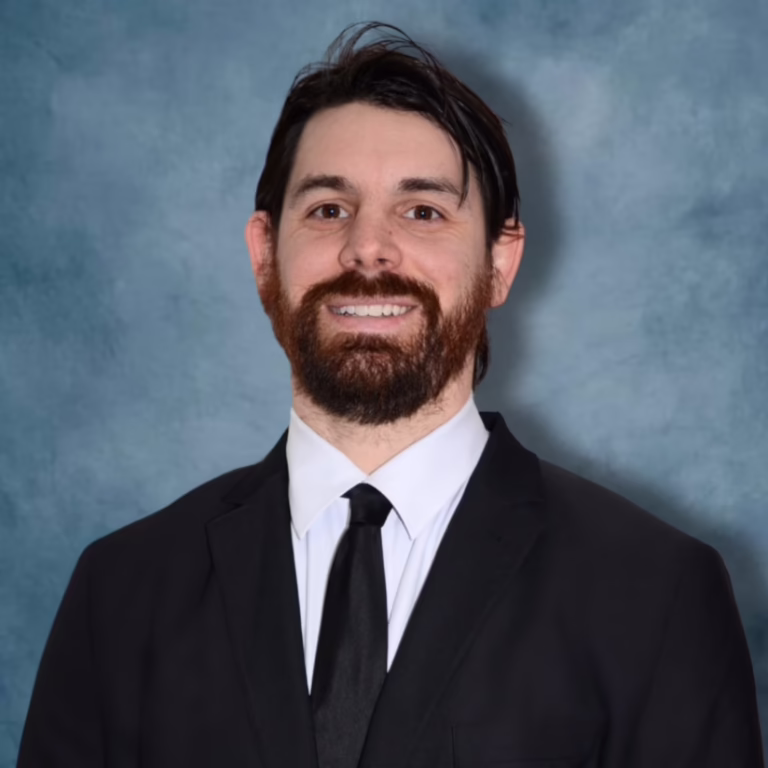 Professional headshot of a man with dark hair and a beard in a black suit and tie, against a blue backdrop.