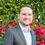 Professional headshot: man in a gray blazer and light blue shirt, smiling with a green garden and pink flowers behind him.
