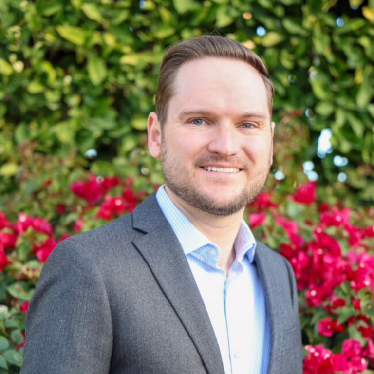 Professional headshot: man in a gray blazer and light blue shirt, smiling with a green garden and pink flowers behind him.