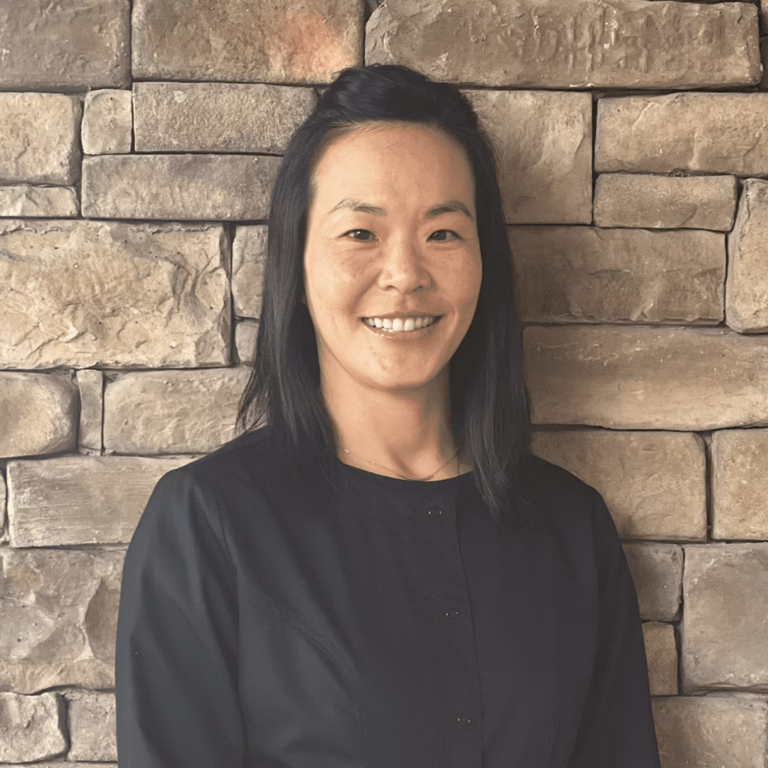 Smiling Asian woman with shoulder-length black hair in a black blouse, standing against a stone wall backdrop.