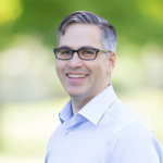 Smiling man with glasses outdoors, light blue shirt, blurred green background
