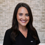 Smiling woman with dark hair in black scrubs posing for a professional headshot against a light brick wall backdrop