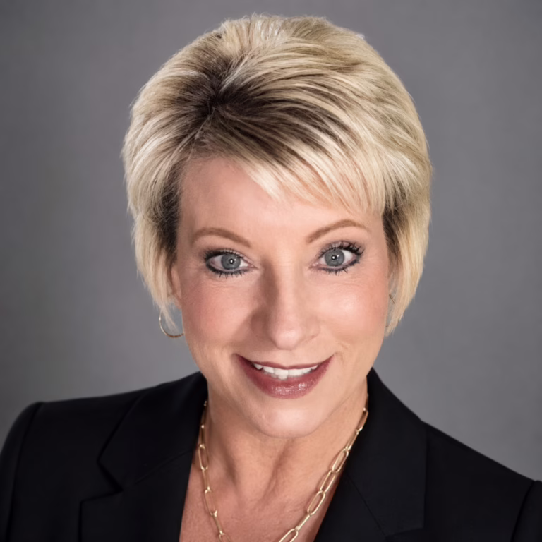Professional headshot of a smiling blonde woman in a black blazer against a gray backdrop, wearing a gold chain necklace.