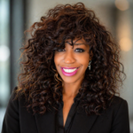 Smiling woman with dark curly hair and pink lipstick wearing a black blazer, standing indoors.