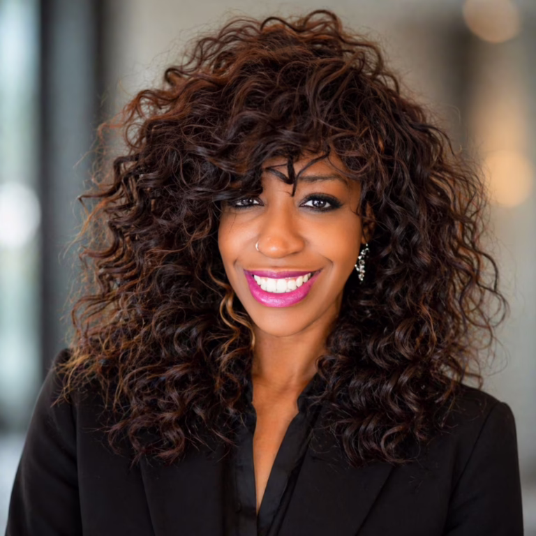 Smiling woman with dark curly hair and pink lipstick wearing a black blazer, standing indoors.