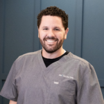 Smiling man in gray scrubs standing in front of blue cabinets in a clinical setting.📍