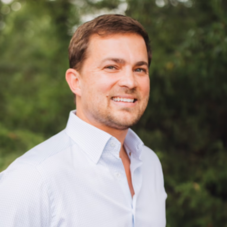 Smiling man in a light blue button-down shirt outdoors with greenery in the background.