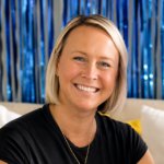 Smiling blonde woman in a black shirt, seated indoors with blue metallic streamers in the background.