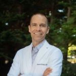 Smiling male doctor in a white coat standing outdoors with trees in the background, arms crossed.