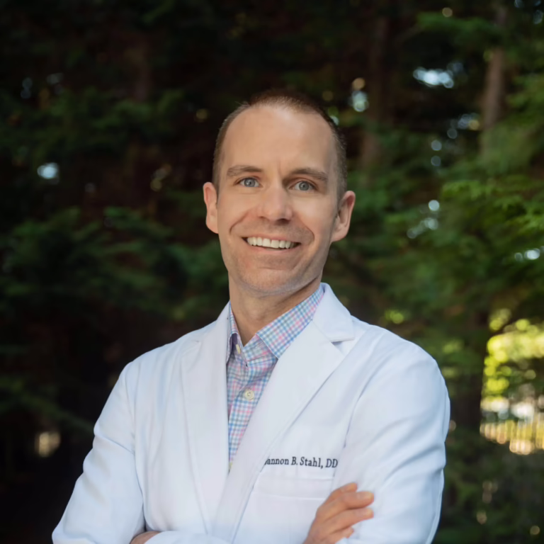 Smiling male doctor in a white coat standing outdoors with trees in the background, arms crossed.
