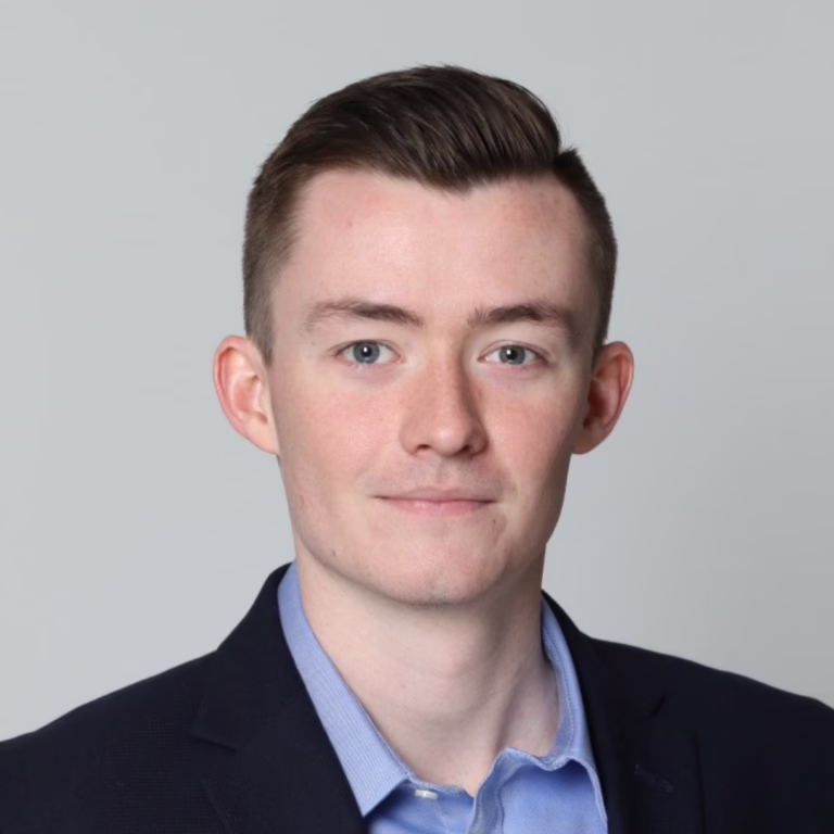 Head-and-shoulders portrait of a young man in a navy blazer and light blue shirt against a gray background.