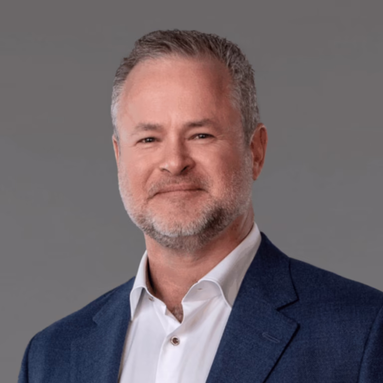 Head-and-shoulders portrait of a middle-aged man with salt-and-pepper hair and a short beard, in a white shirt and blue blazer against a gray backdrop.