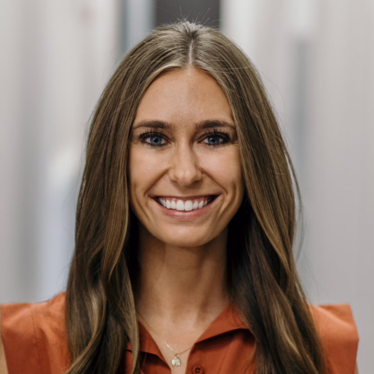 Smiling woman with long brown hair wearing an orange blouse, neutral office background.