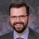 Smiling man with glasses and a beard in a dark suit, lavender shirt, and purple tie, posing for a professional headshot against a neutral backdrop.