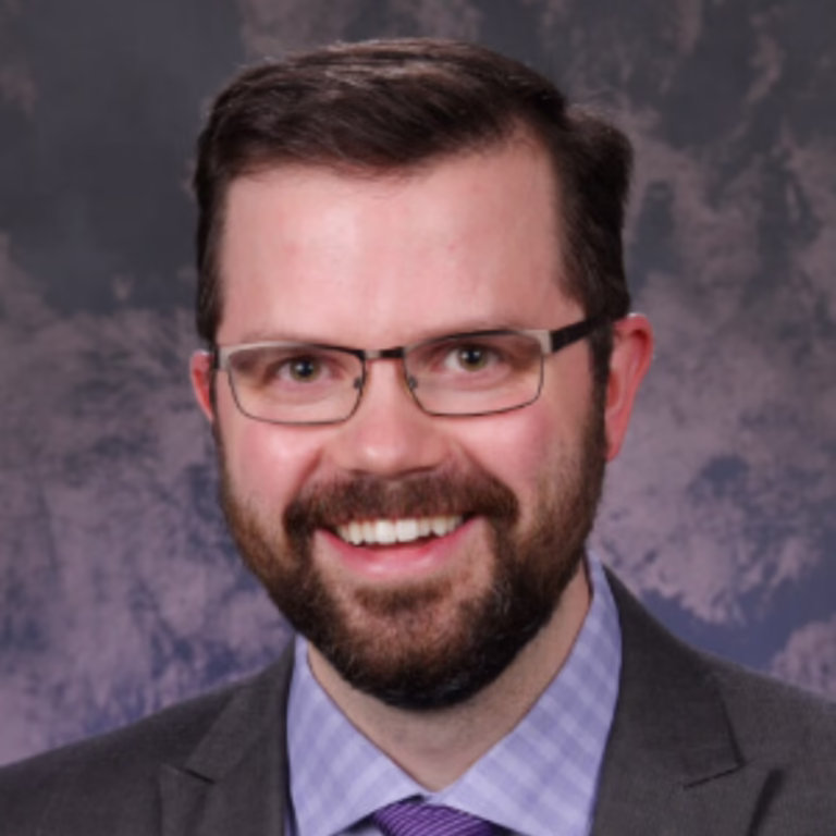 Smiling man with glasses and a beard in a dark suit, lavender shirt, and purple tie, posing for a professional headshot against a neutral backdrop.