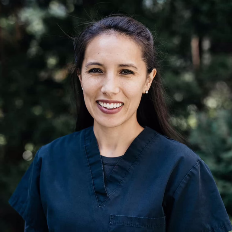 Smiling woman in navy scrubs outdoors with green trees behind her.