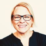 Smiling woman with short blond hair and black-rimmed glasses looking at the camera, wearing a dark top.
