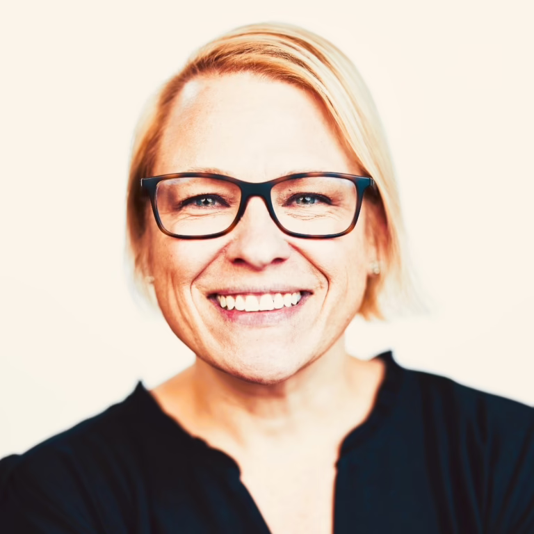 Smiling woman with short blond hair and black-rimmed glasses looking at the camera, wearing a dark top.